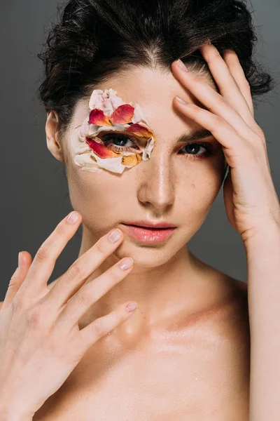 Hermosa joven posando con pétalos florales alrededor del ojo aislado en gris - foto de stock