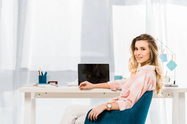 Jolie jeune femme en chemise à carreaux assis sur le lieu de travail et regardant la caméra — Photo de stock