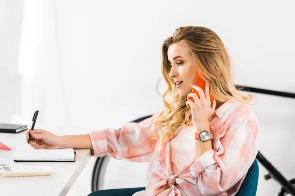 Pretty young woman talking on smartphone and writing in notebook at workplace — Stock Photo
