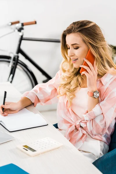 Чарівна жінка в рожевій сорочці говорить на смартфоні і пише в блокноті в домашньому офісі — стокове фото
