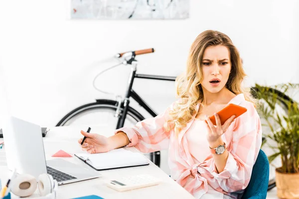 Mujer rubia irritada con pluma en la mano mirando el teléfono inteligente en el lugar de trabajo - foto de stock