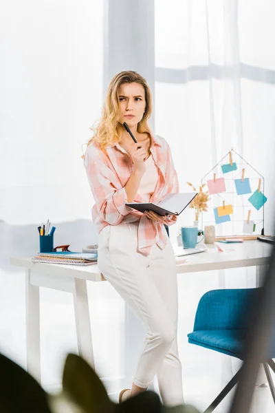 Mujer pensativa de pie en el lugar de trabajo y la celebración de cuaderno con pluma - foto de stock