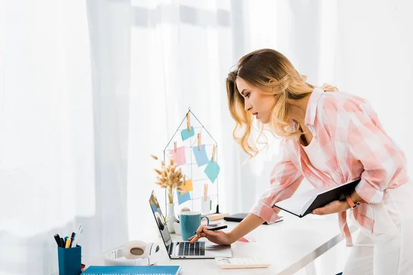 Concentrated young woman with notebook using laptop at home office — Stock Photo