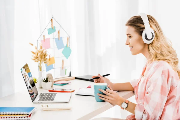 Side view of smiling woman listening music and drinking coffee at workplace — Stock Photo