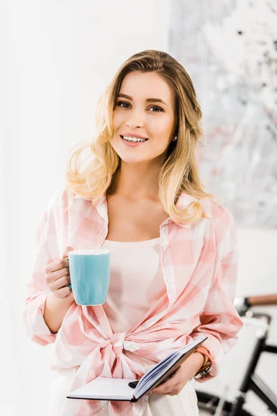 Jolie femme blonde en chemise à carreaux tenant tasse de café et carnet — Photo de stock