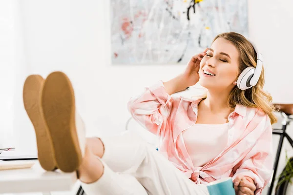 Smiling young woman listening music with closed eyes and legs on table — Stock Photo