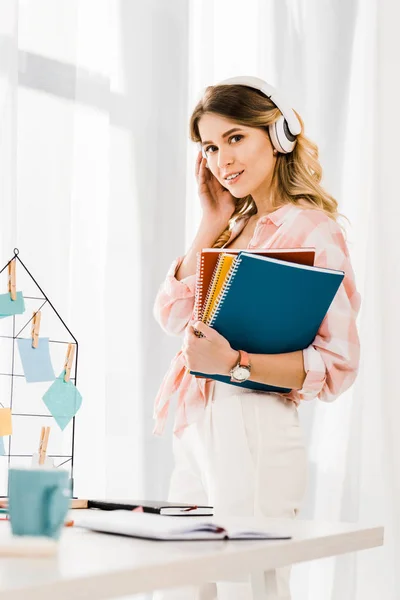 Curiosa mujer bonita con cuadernos escuchando música y mirando a la cámara - foto de stock