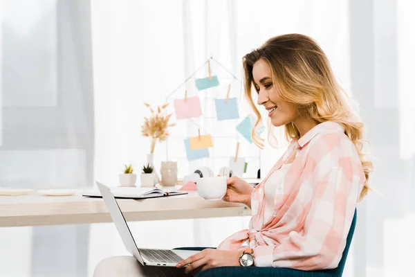Fröhliche junge Frau im karierten Hemd, die Kaffee trinkt und Laptop benutzt — Stockfoto