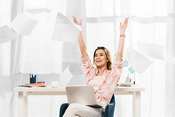 Mulher rindo com laptop jogando fora documentos no local de trabalho — Fotografia de Stock