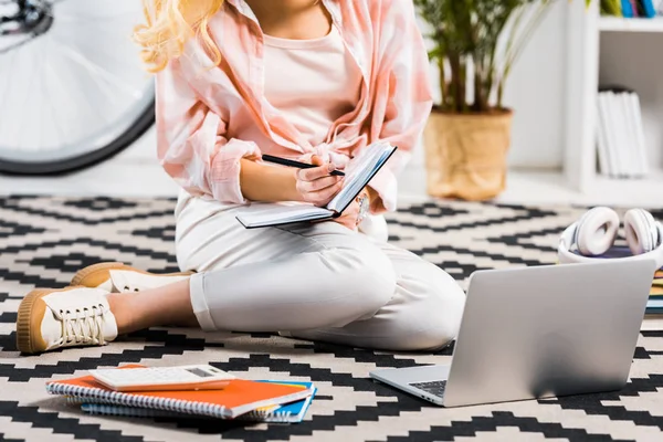 Partial view of woman with laptop sitting on carpet and writing on notebook — Stock Photo