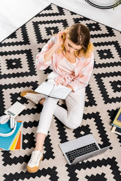 Geschäftiges Mädchen in weißen Hosen auf Teppich sitzend und Notizbuch in der Hand — Stockfoto