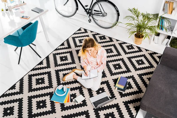 Frau sitzt auf Teppich und schreibt zu Hause in Notizbuch — Stockfoto