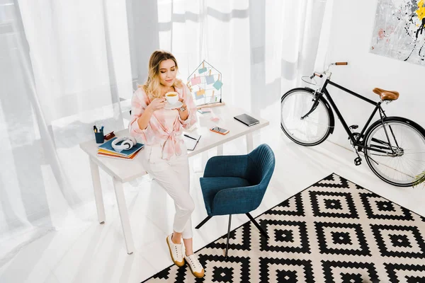 Mujer rubia bonita en pantalones blancos bebiendo café en casa oficina - foto de stock