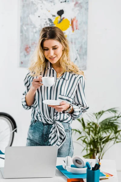 Hermosa chica con camisa a rayas bebiendo café y mirando el portátil - foto de stock