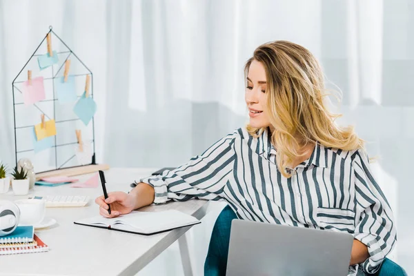 Mujer sonriente con camisa a rayas sosteniendo laptop y escribiendo en cuaderno - foto de stock