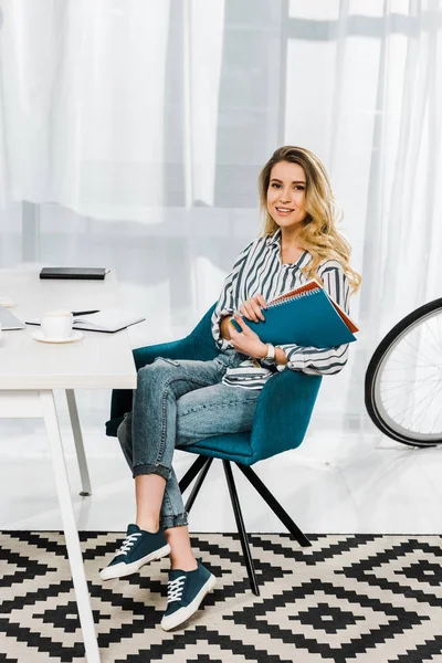 Charmante blonde Frau sitzt im Sessel und hält zu Hause im Büro Notizbücher — Stockfoto