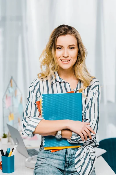 Lieto giovane donna in camicia a righe che tiene i quaderni e guarda la fotocamera con sorriso — Foto stock