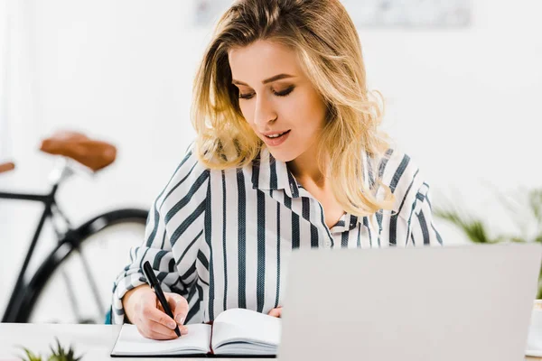 Charmante blonde Frau in gestreiftem Hemd schreibt in Notizbuch am Arbeitsplatz — Stockfoto