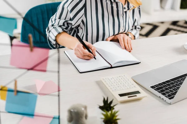 Vista parcial de la mujer con camisa a rayas escribiendo en cuaderno en el lugar de trabajo - foto de stock