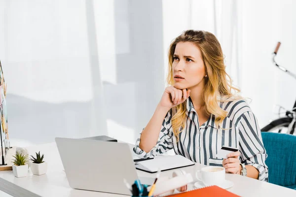 Pensive woman in striped shirt holding credit card at workplace — Stock Photo