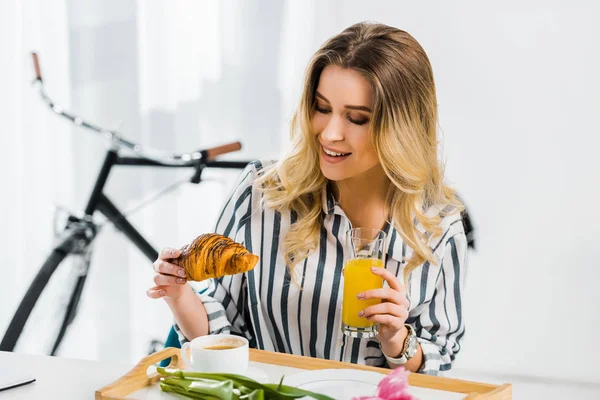 Fröhliche Frau im gestreiften Hemd isst Croissant und trinkt Orangensaft — Stockfoto