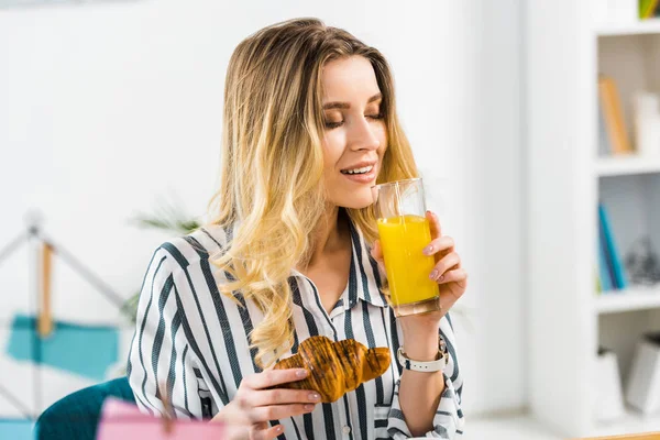 Relaxed woman in striped shirt holding croissant and drinking orange juice — Stock Photo