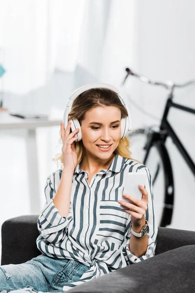 Mujer alegre con camisa a rayas sosteniendo smartphone y escuchando música en auriculares - foto de stock