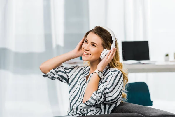 Glückliche Frau im gestreiften Hemd auf Sofa sitzend und Musik über Kopfhörer hörend — Stockfoto