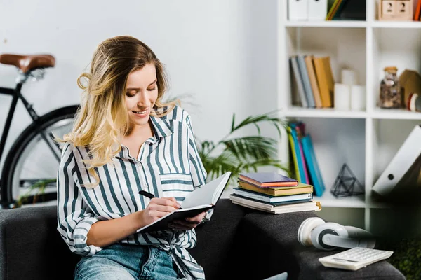 Freudige junge Frau in gestreiftem Hemd sitzt auf Sofa und schreibt in Notizbuch — Stockfoto