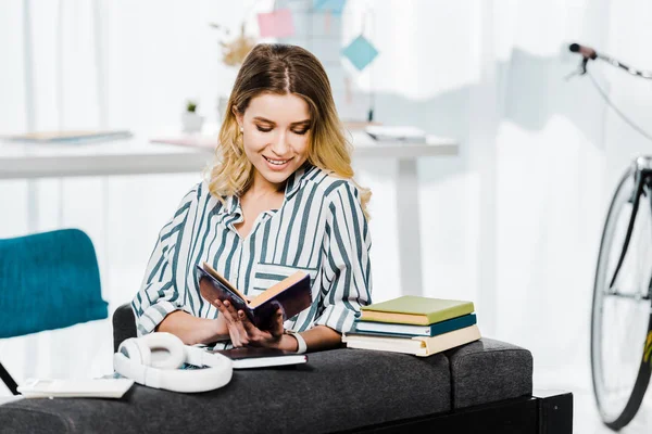 Menina sorridente em camisa listrada sentado no sofá e livro de leitura — Fotografia de Stock