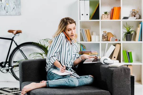 Frau im gestreiften Hemd spricht auf Smartphone und benutzt Laptop zu Hause — Stockfoto