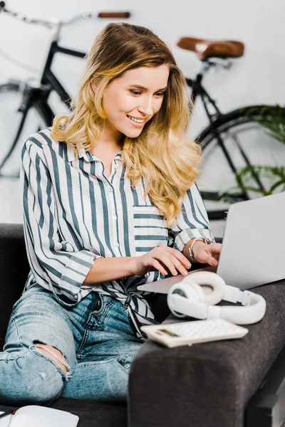 Радостная женщина в полосатой рубашке сидит на диване и печатает на клавиатуре ноутбука — стоковое фото