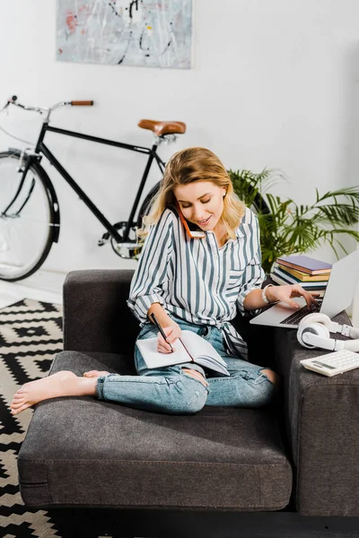 Woman with laptop talking on smartphone and writing in notebook — Stock Photo