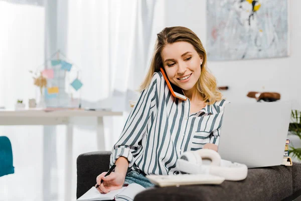 Lächelnde junge Frau spricht auf Smartphone und schaut auf Laptop — Stockfoto