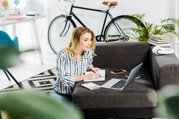 Woman in striped shirt writing in notebook and looking at laptop — Stock Photo