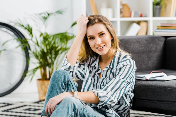 Joyeux jeune femme en chemise rayée assise sur le tapis à la maison — Photo de stock