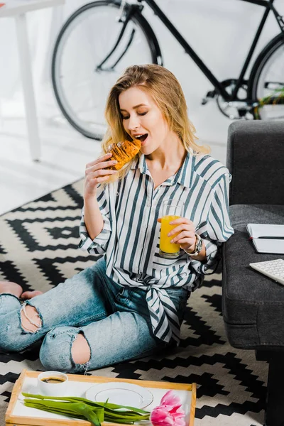 Блондинка в смугастій сорочці їсть круасан і тримає склянку апельсинового соку — стокове фото