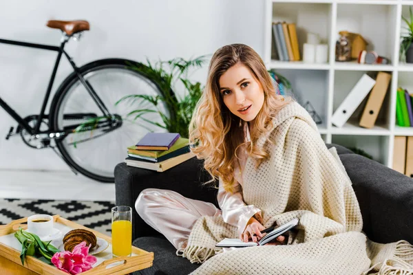 Atemberaubendes Mädchen mit Strickkarierten sitzt auf dem Sofa und hält Notizbuch — Stockfoto