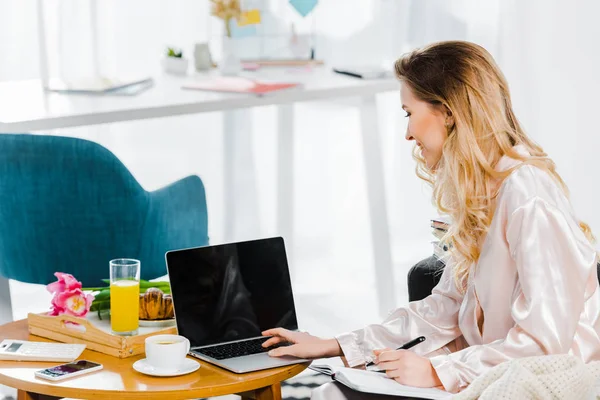 Jeune femme en pyjama satiné utilisant un ordinateur portable avec écran blanc pendant le petit déjeuner — Photo de stock