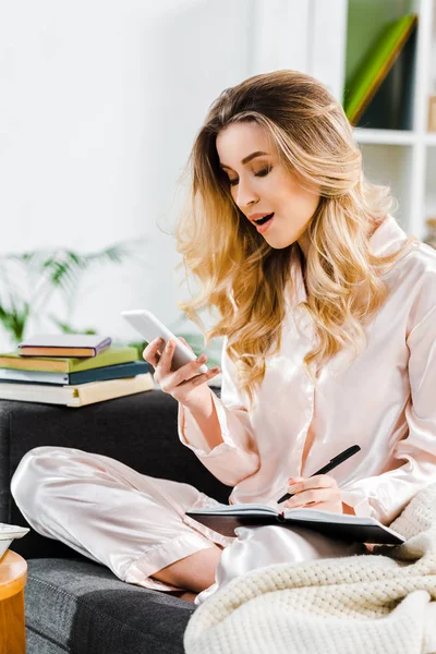 Blondine im Schlafanzug sitzt mit Notizbuch auf Sofa und hält Smartphone in der Hand — Stockfoto