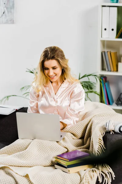 Chica sonriente en pijama con cuadros sentados en el sofá y el uso de la computadora portátil - foto de stock
