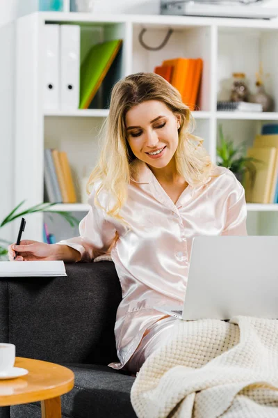 Jolie femme en pyjama utilisant un ordinateur portable et écrivant dans un carnet à la maison — Photo de stock
