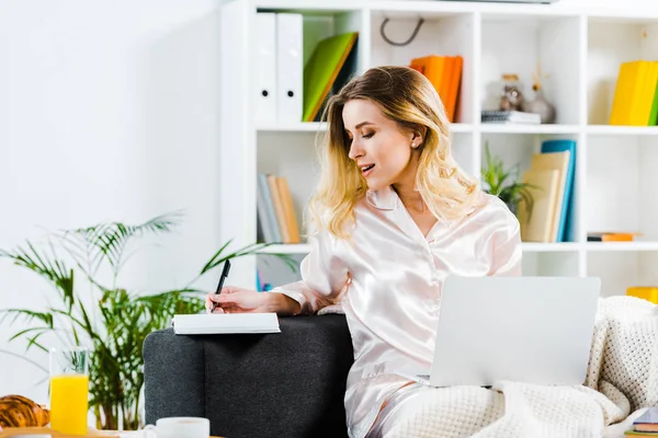 Affascinante donna in pigiama seduta sul divano con laptop e scrittura su taccuino — Foto stock