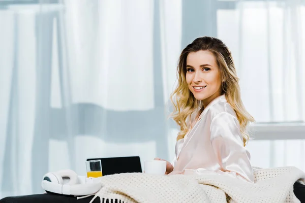 Charming young woman in satin pyjamas holding coffee cup and smiling at camera — Stock Photo