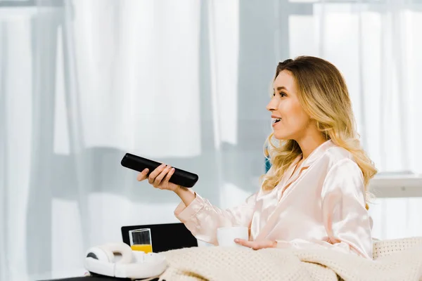 Smiling woman in pyjamas holding coffee cup and tv remote control — Stock Photo