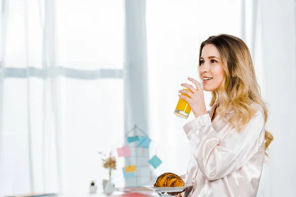 Chica inspirada en pijamas sosteniendo croissant y bebiendo jugo de naranja en la mañana - foto de stock