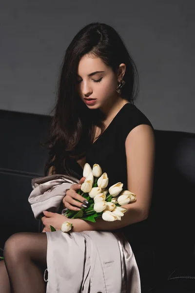 Attractive brunette woman in black dress sitting with tulips on dark background — Stock Photo