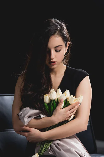 Attractive brunette woman in dress sitting on couch with white tulips on black background — Stock Photo