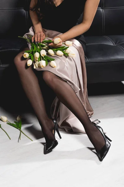 Обрезанный вид женщины в черном платье, держащей белые тюльпаны и сидящей на темном диване — стоковое фото