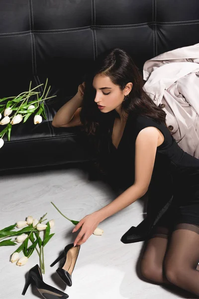 Attraktive Brünette im schwarzen Kleid auf dem Boden mit weißen Tulpen in der Nähe der dunklen Couch — Stockfoto